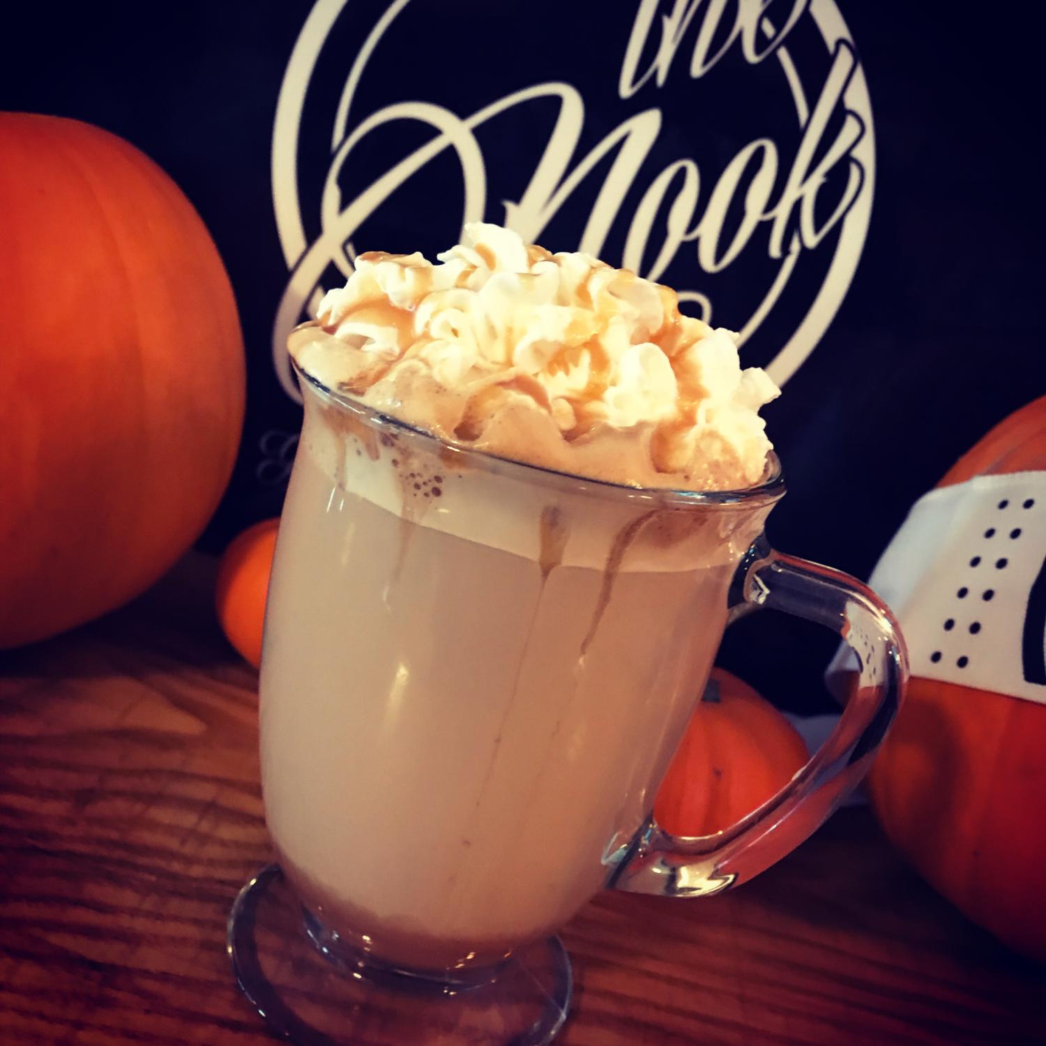 A glass mug of hot chocolate with whipped cream, in front of The Nook logo and pumpkins
